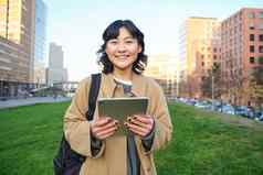 快乐亚洲女孩站街大学学生走数字平板电脑手微笑站城市中心