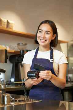 肖像微笑亚洲咖啡师咖啡商店员工回来终端信贷卡帮助客户端支付非接触式咖啡馆