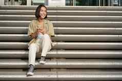 肖像微笑亚洲女孩坐在楼梯智能手机浏览互联网移动电话休息在户外城市中心