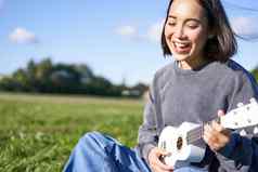 快乐人爱好微笑亚洲女孩玩尤克里里琴吉他唱歌坐着公园在户外毯子