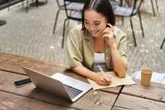 肖像年轻的女人研究在线坐着移动PC写作使笔记电脑屏幕坐着咖啡馆在户外