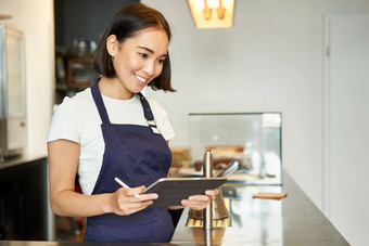 小咖啡馆业务微笑亚洲女孩咖啡师围裙平板电脑回来终端处理咖啡订单计数器图片