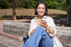 快乐微笑亚洲女孩坐着板凳上公园浏览社会媒体持有智能手机移动电话应用程序
