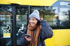 肖像女孩站公共汽车停止等待公共运输检查时间表智能手机应用程序持有移动电话穿温暖的衣服
