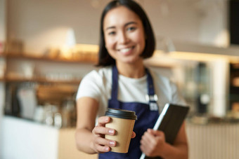 模糊拍摄年轻的微笑咖啡师女孩给外卖杯咖啡带外卖卡布奇诺咖啡快乐工作咖啡馆