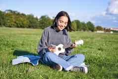 微笑亚洲女孩吉他应用程序唱歌玩尤克里里琴窥视和弦智能手机坐着在户外公园毯子