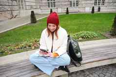 年轻的红色头发的人女孩学生坐在板凳上智能手机应用程序手表视频在线发送消息移动电话