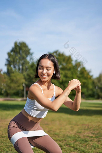 垂直拍摄年轻的适合女人下蹲公园伸展运动乐队腿微笑很高兴锻炼