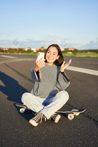 快乐青少年亚洲女孩需要自拍视频聊天智能手机应用程序坐在滑板溜冰者记录视频博客坐在冲浪老手