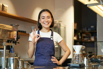 肖像微笑朝鲜文咖啡师女孩计数器穿蓝色的围裙作品咖啡商店显示标志