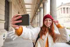 可爱的年轻的红色头发的人女人需要自拍街移动电话使照片智能手机应用程序街