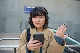 肖像亚洲女孩耳机复杂的智能手机屏幕困惑文本消息通知站街耸了耸肩