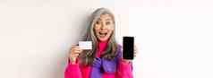 在线购物特写镜头惊讶亚洲高级女人显示空白移动屏幕塑料信贷卡快乐站白色背景