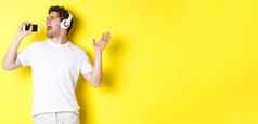 快乐的家伙玩卡拉ok应用程序耳机唱歌智能手机麦克风站黄色的背景