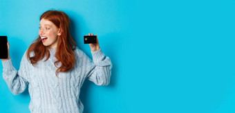 兴奋红色头发的人女孩显示移动电话屏幕信贷卡展示在线商店应用程序站蓝色的背景