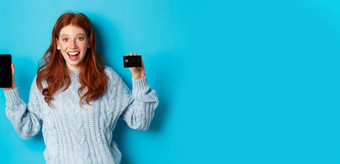兴奋红色头发的人女孩显示移动电话屏幕信贷卡展示在线<strong>商店应用</strong>程序站蓝色的背景