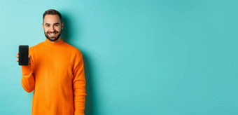 英俊的有胡子的的家伙橙色毛衣显示智能手机屏幕微笑显示促销在线绿松石背景