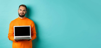 英俊的有胡子的男人。橙色毛衣显示移动PC屏幕展示促销扮鬼脸失望心烦意乱站光蓝色的背景