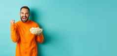 快乐的英俊的男人。橙色毛衣欢呼欢乐吃爆米花看体育使拳头泵满意站蓝色的背景