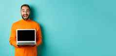 英俊的有胡子的男人。橙色毛衣显示移动PC屏幕展示促销站光蓝色的背景