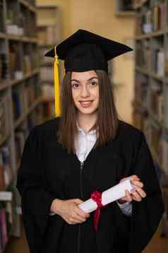 快乐年轻的女人研究生礼服持有文凭图书馆
