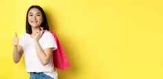 快乐的亚洲女购物者逗乐持有购物袋显示拇指推荐商店折扣站黄色的背景