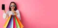 有吸引力的亚洲女人显示智能手机应用程序购物袋购买在线应用程序站粉红色的背景