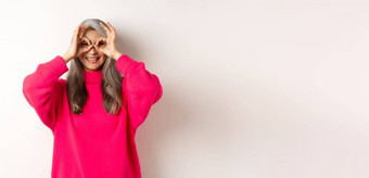快乐有趣的亚洲高级女人使手双筒望远镜手指微笑站粉红色的毛衣白色背景