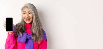 在线购物时尚的美丽的亚洲高级女人灰色头发显示空白智能手机屏幕很高兴站白色背景