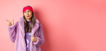 时尚购物概念持怀疑态度中年亚洲女人紫色的皮毛外套指出手指左不高兴伤心表达式站粉红色的背景