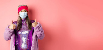 冠状病毒购物概念时尚的亚洲祖母呼吸器闪闪发光的紫色的衣服人造皮毛外套显示竖起大拇指推荐商店脸面具