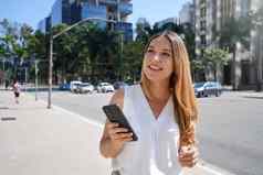 美丽的年轻的女人持有智能手机走金融区肖像华丽的微笑女移动电话现代大都市