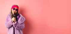 时尚概念时尚的高级亚洲女人唱歌卡拉ok执行阶段麦克风穿时尚的太阳镜紫色的人造皮毛外套粉红色的背景