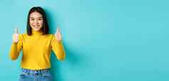 快乐的亚洲女模型显示拇指手势微笑深刻的印象赞美好产品站蓝色的背景