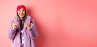 时尚购物概念很酷的亚洲高级女人时尚的假的皮毛外套指出手指相机检查促销提供站粉红色的背景