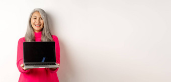 电子商务概念美丽的亚洲高级女商人显示空白移动PC屏幕微笑站粉红色的毛衣白色背景