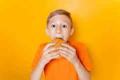 男孩橙色t恤持有汉堡手贪婪地咬黄色的背景
