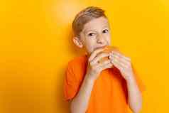 男孩橙色t恤持有汉堡手贪婪地咬黄色的背景