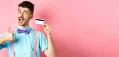 购物概念节日的家伙蝴蝶结眨眼相机显示拇指塑料信贷卡推荐好商店提供促进银行站粉红色的背景