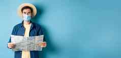 科维德流感大流行旅行概念快乐旅游夏天假期穿医疗面具稻草他持有路地图站蓝色的背景