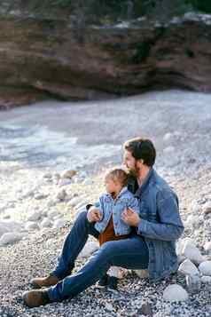 爸爸坐在石头拥抱女孩卵石海滩背景岩石女儿爸爸海一边视图