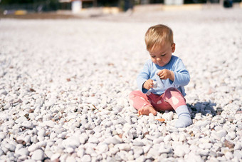 可爱的婴儿蓝色的上衣红色的裤子坐在卵石海滩鞠躬头持有鹅卵石手
