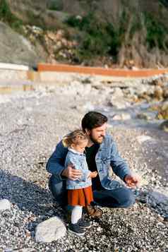 微笑爸爸蹲女孩将鹅卵石岩石海滩水特写镜头