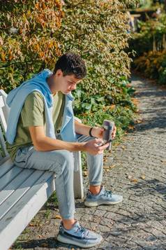 少年坐在板凳上秋天公园饮料咖啡热杯子电话肖像英俊的快乐的的家伙坐着板凳上新鲜的空气设备浏览媒体smm喝拿铁城市户外