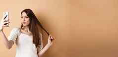 时尚的年轻的女人玩头发采取自拍智能手机使照片社会媒体站米色背景