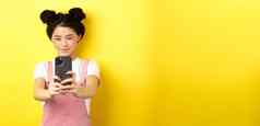 时尚的亚洲女孩采取照片智能手机使视频手机微笑站黄色的背景