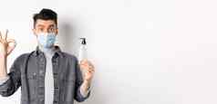 健康的人科维德概念兴奋男人。医疗面具持有瓶好手洗手液显示标志推荐防腐剂站白色背景