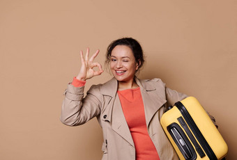 快乐的女人旅行者乘客黄色的手提箱显示标志眨眼微笑相<strong>机米</strong>色背景
