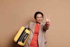 可爱的少数民族女人乘客携带手提箱微笑手势和平标志米色孤立的背景