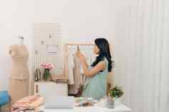 美丽的年轻的亚洲时尚设计师采取照片工作智能手机女装裁缝工作室